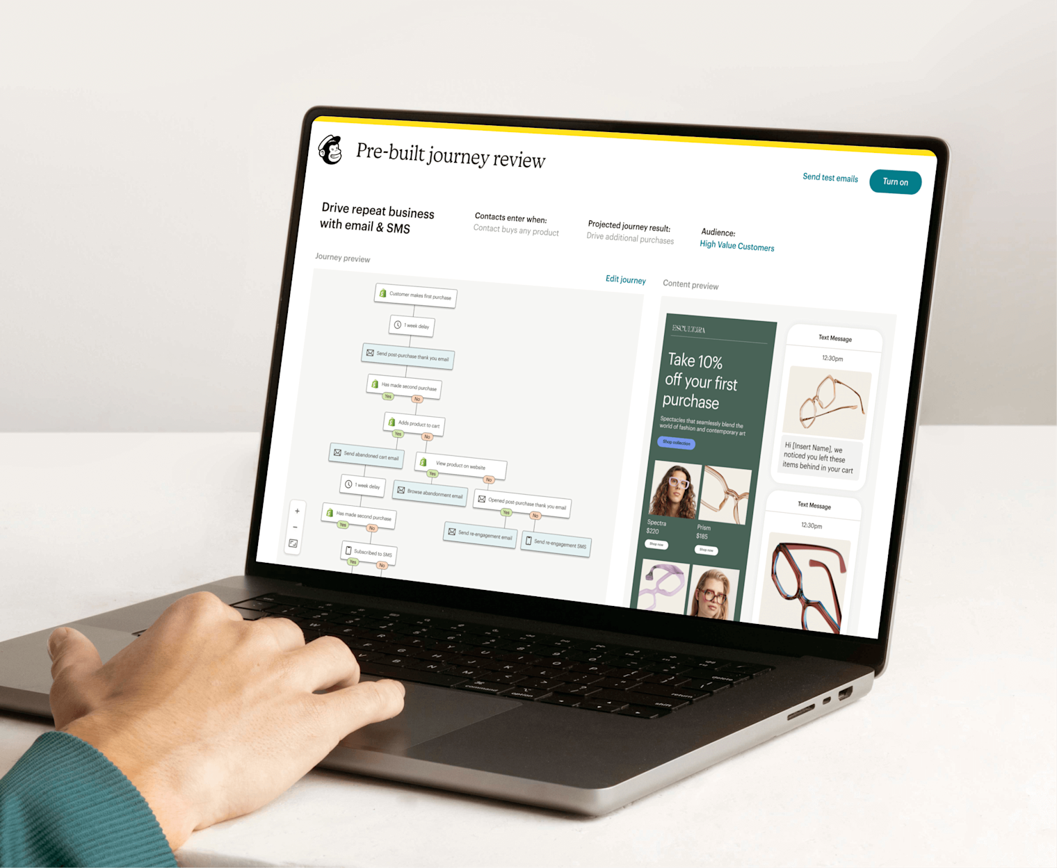 Image of a person sitting at a desk, looking at a laptop screen displaying Mailchimp’s Pre-built journey review feature. The feature shows a preview of an automation journey and an abstract view of email and SMS interfaces from the fictitious brand Escultura.